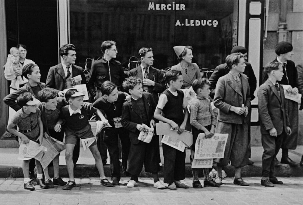 A crowd gathered in front of Mr. Pierre Cloarec's bicycle shop, who is racing in the Tour de France. Pleybon, France, 1939 © Robert Capa © International Center of Photography / Magnum Photos